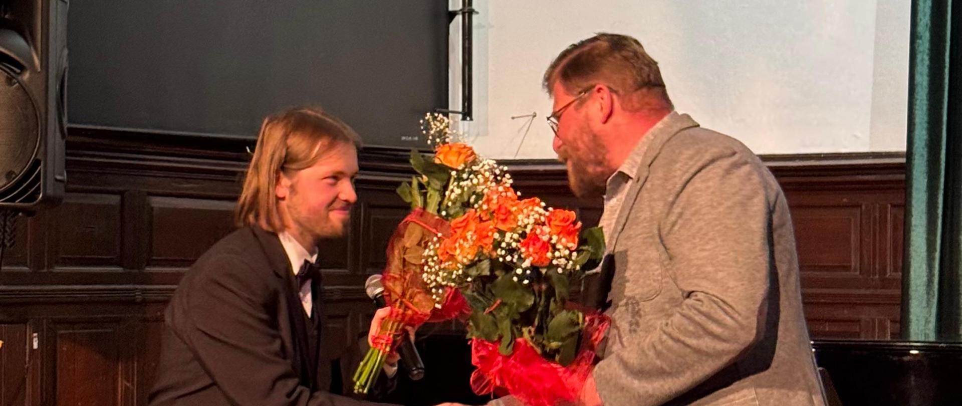 Dwóch mężczyzn w eleganckich strojach, Młodszy mężczyzna przyjmuje od starszego kwiaty i gratulacje.. W tle znajduje się fortepian. 