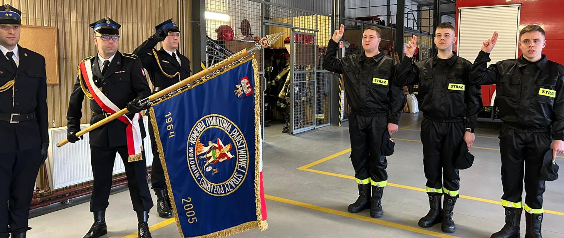 Zdjęcie przedstawia poczet sztandarowy oraz trzech strażaków składających ślubowanie. Chorąży pochyla sztandar do przodu, pozostali członkowie pocztu oddają honor. Ślubujący strażacy trzymają uniesione do góry prawe ręce. Składają ślubowanie.