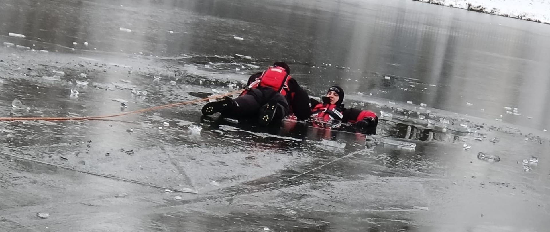 Strażak w sprzęcie asekuracyjnym oraz zabezpieczony linką czołga się po odzie do pozoranta udającego osobę, pod którą załamał się lód.