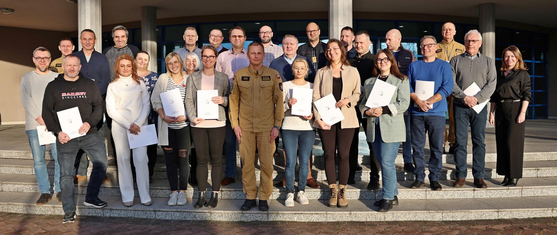 Pamiątkowe zdjęcie grupowe uczestników szkolenia inspektorów ochrony przeciwpożarowej (23 osoby) w towarzystwie Zastępcy Komendanta Centralnej Szkoły PSP, kobiety oraz dwóch funkcjonariuszy CS PSP