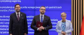 Na tle wielkiego niebieskiego ekranu stoją wiceminister Szeptycki, minister Kulasek i wiceminister Mrówczyńska.