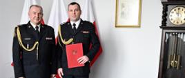 Dwóch strażaków w mundurach wyjściowych ze sznurem stoi obok siebie jeden z nich trzyma czerwoną teczkę za nimi są trzy flagi Polski na ścianie wisi obraz w ścianie są wyłączniki obok stoi zegar. 