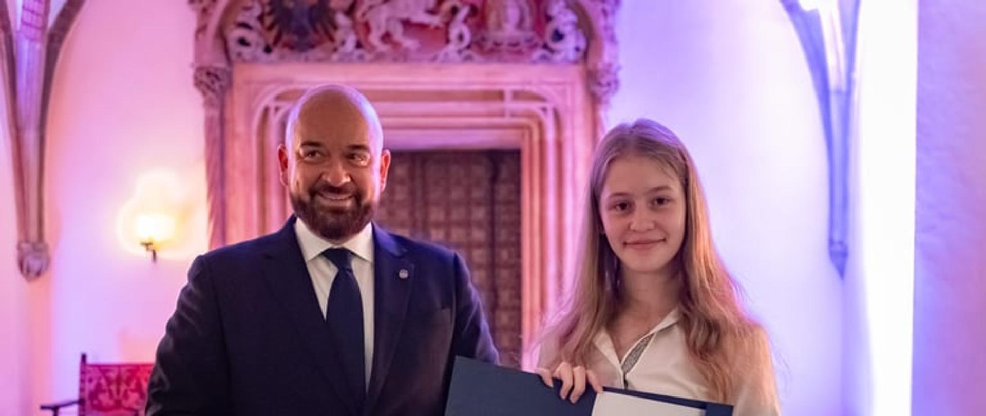 Mężczyzna ubrany w ciemny garnitur, obok stoi dziewczyna ubrana w białą bluzkę i czarną spódnicę. Dziewczyna w rękach trzyma teczkę z dyplomem.