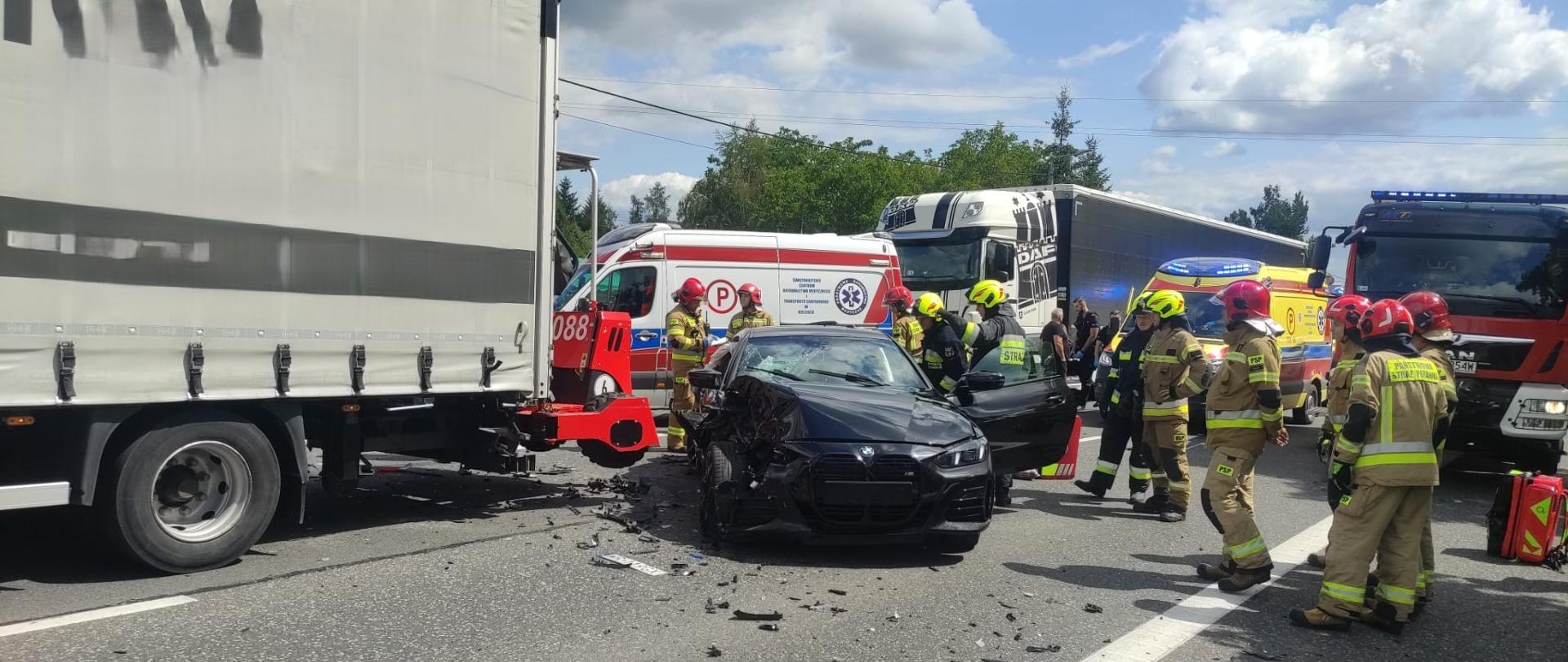 Zdjęcie przedstawia miejsce wypadku: w centrum obrazu zniszczony samochód osobowy a za nim ambulans, z lewej ciężarówka biorąca udział w zdarzeniu. Po prawej widać samochód strażacki, kolejny ambulans i drugą z biorących udział w zderzeniu ciężarówkę. Między pojazdami stoją strażacy.
