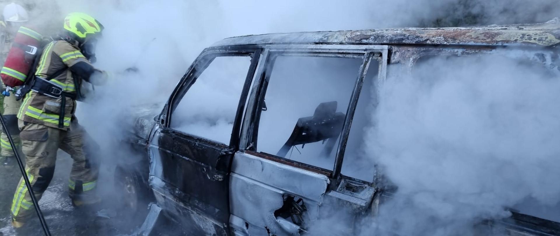Wrak samochodu, z którego unoszą się kłęby dymu oraz pary wodnej. Obok strażak gasi samochód.