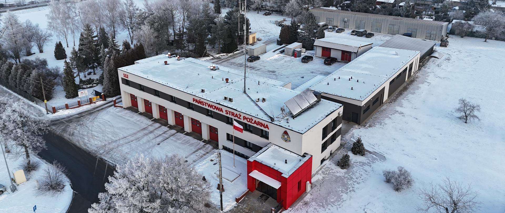 Zdjęcie lotnicze przedstawiające siedzibę Komendy Powiatowej Państwowej Straży Pożarnej w Zduńskiej Woli zimą. Na pierwszym planie widoczny jest nowoczesny budynek strażnicy z jasną elewacją i czerwonymi bramami garażowymi, nad którymi znajduje się napis »Państwowa Straż Pożarna«. Obok budynku wznosi się wysoki maszt antenowy. Teren wokół pokryty jest śniegiem, a w tle widać zabudowania miasta, drzewa oraz obiekty przemysłowe.
