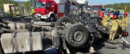 „Wrak rozbitego samochodu ciężarowego, przy którym strażacy uwalniają zakleszczonego kierowcę. W tle widoczne są samochody pożarnicze, radiowozy oraz pojazdy stojące w korku.