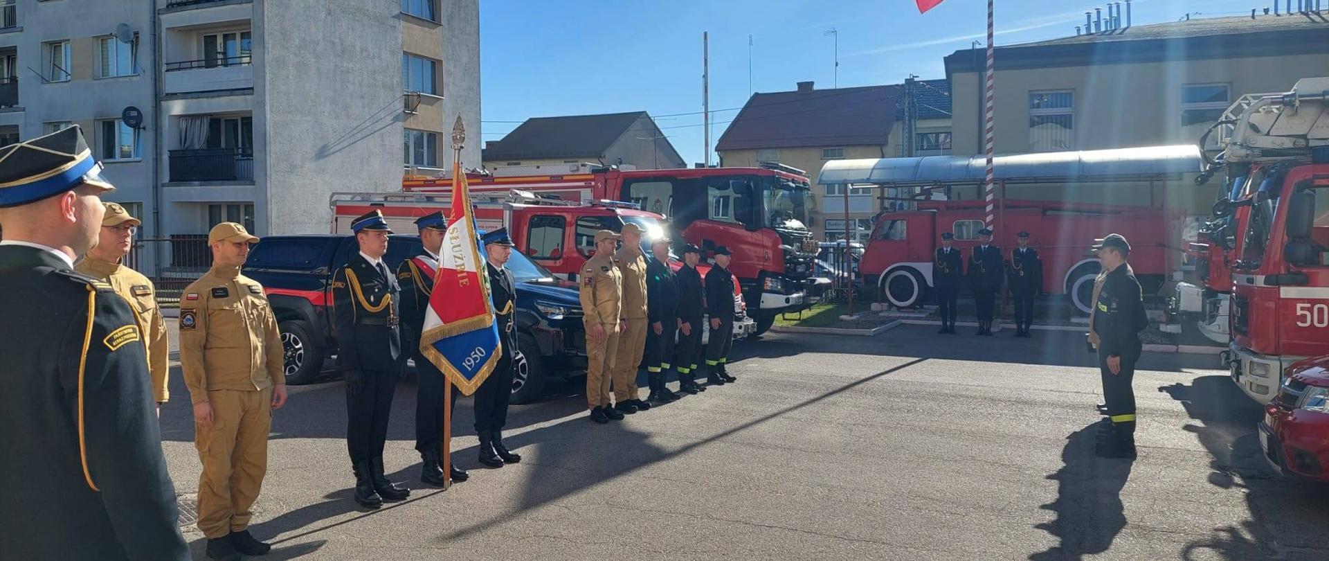 Funkcjonariusze PSP stoją w dwóch rzędach. Widać również wozy strażackie i poczet flagowy. 
