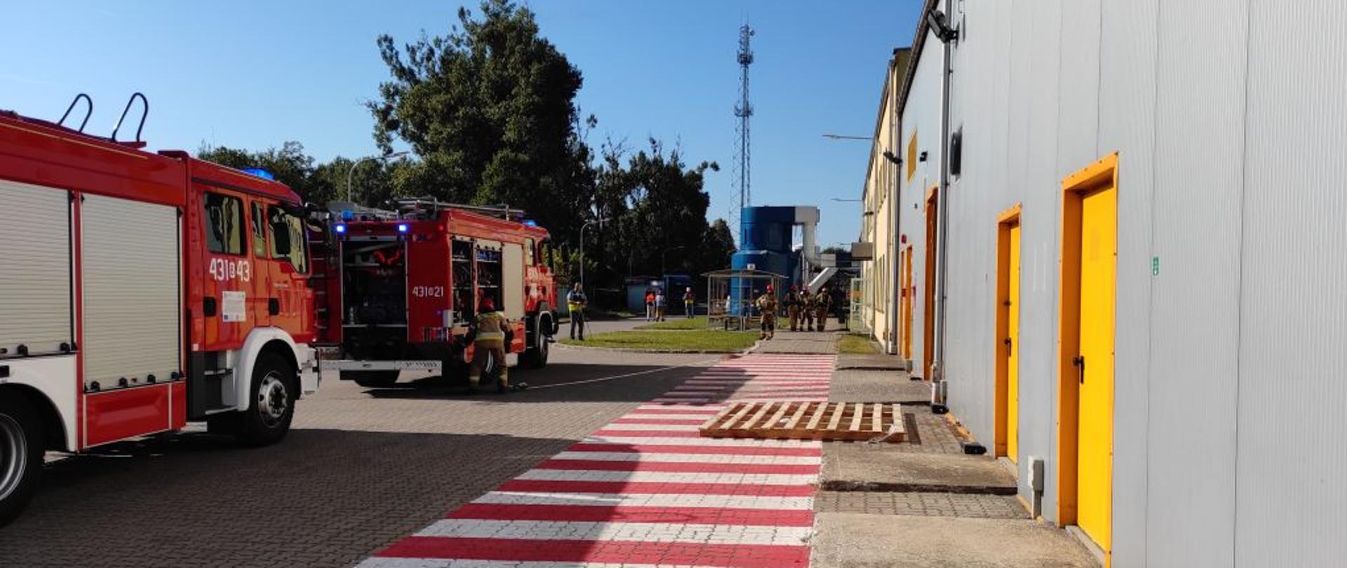 Po prawej stronie szary budynek produkcyjny z żółtymi drzwiami. Po środku na drodze namalowane białoczerwone pasy. Po lewej dwa strażackie samochody w okól których strażacy rozwijają węże.