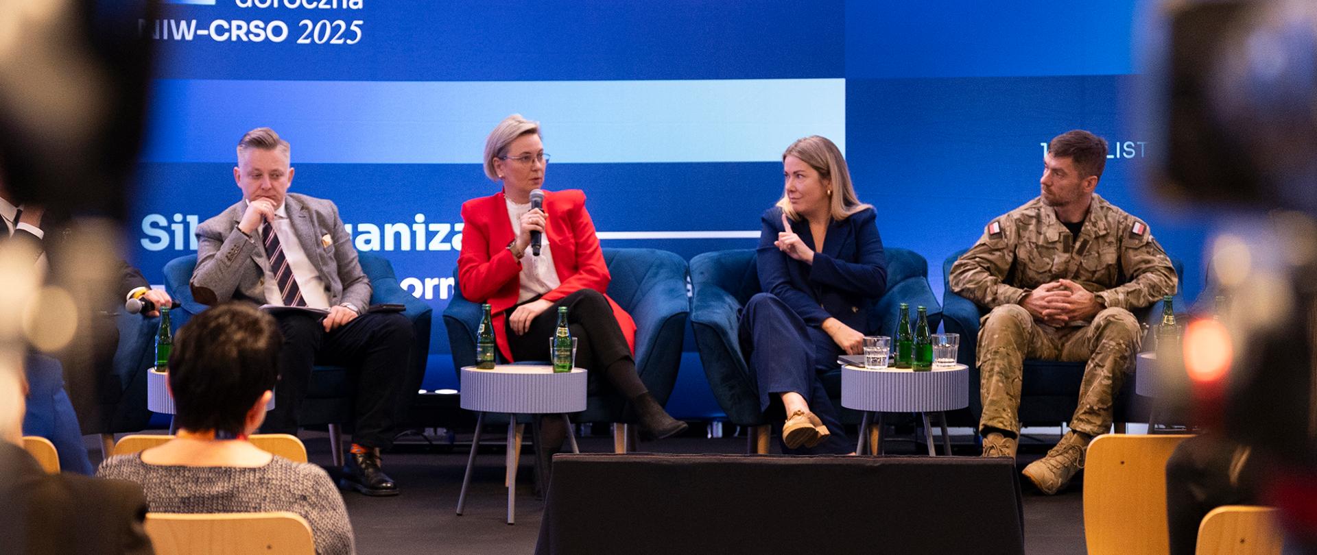 Na zdjęciu uczestnicy podczas debaty głównej. Prelegenci zajmują fotele na scenie, ustawione w rzędzie jeden obok drugiego. Od lewej widoczni: Minister Jakub Stefaniak, Sekretarz Stanu Adriana Porowska, Sekretarz Stanu Magdalena Roguska, Podpułkownik Paweł Wronka. Sekretarz Stanu Adriana Porowska mówi do mikrofonu, który trzyma w dłoni. W tle ekran, na którym wyświetlona jest grafika tytułowa konferencji. Na pierwszym planie widoczne są sylwetki osób siedzących na widowni oraz, po bokach kadru, rozmyte kształty kamer telewizyjnych na statywach.