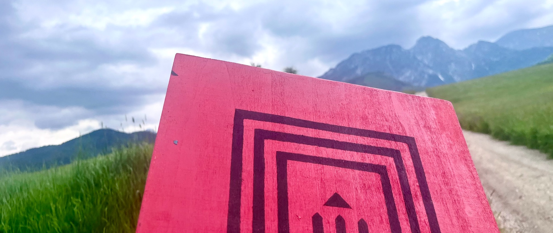 Na zdjęciu widać:
Czerwoną tablicę lub znak ustawiony na pierwszym planie. Znajduje się na nim czarny, geometryczny symbol przypominający labirynt lub stylizowaną bramę z trzema pionowymi liniami zakończonymi strzałką skierowaną w górę.
Kamienistą drogę lub ścieżkę, biegnącą po prawej stronie kadru.
Zielone łąki i wysoką trawę po obu stronach ścieżki.
Góry w tle, częściowo zamglone lub rozmyte przez głębię ostrości.
Zachmurzone niebo z dużą ilością ciemnych i jasnych chmur, sugerujące zmienną pogodę.