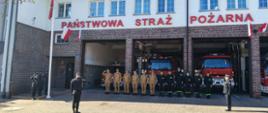 Szeroki kadr przedstawiający fasadę piętrowego budynku Państwowej Straży Pożarnej z dużym czerwonym napisem nad bramami garażowymi. Przed budynkiem, na placu z kostki brukowej, trwa uroczystość. Pośrodku stoi szereg strażaków: część w piaskowych mundurach służbowych, część w czarnych mundurach koszarowych. Przed nimi stoją dwaj dowódcy w ciemnogranatowych mundurach wyjściowych, zwróceni w stronę masztu flagowego po lewej stronie. Na maszt wciągana jest biało-czerwona flaga, a asystuje przy tym poczet flagowy składający się z trzech strażaków. W tle widać czerwone wozy strażackie zaparkowane w garażach.