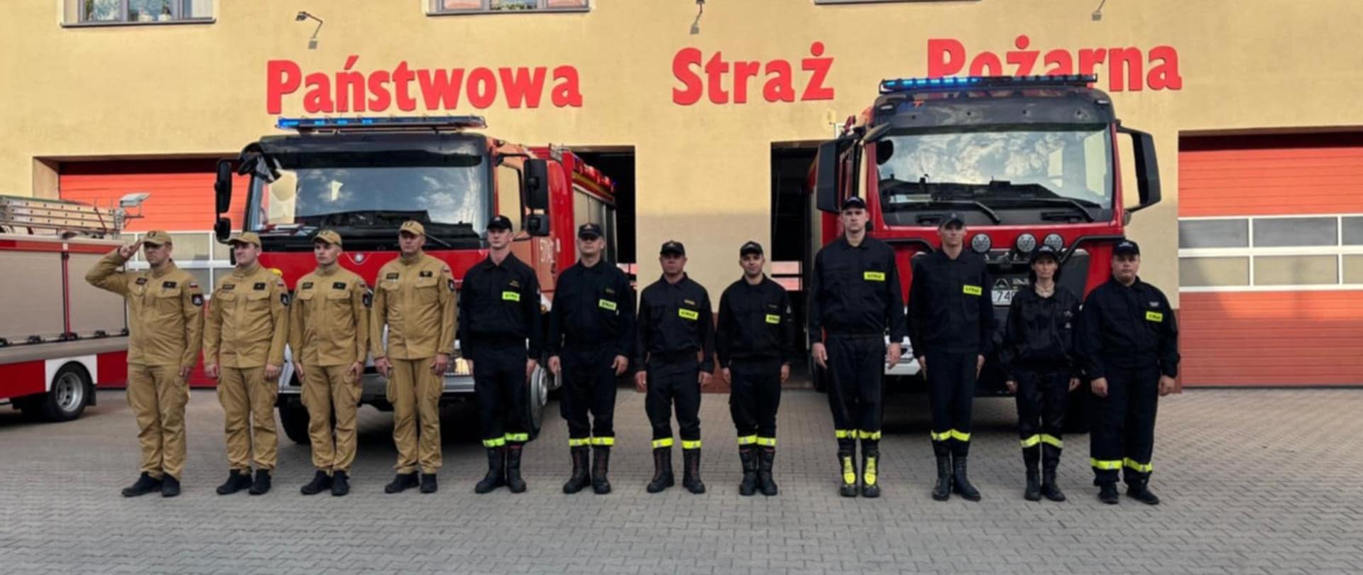 Plac przed komendą. Na tle budynku komendy i wozów bojowych, w szeregu stoją strażacy PSP i OSP, którzy oddają hołd tragicznie zmarłemu strażakowi OSP. Dowódca stojący na prawym skrzydle oddaje honor przez salutowanie. Czterech strażaków ma na sobie ubranie służbowe w kolorze piaskowym, pozostali w ciemnym.