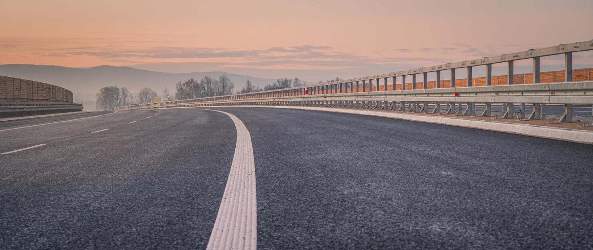Droga ekspresowa prowadzona na obiekcie inżynierskim (najprawdopodobniej wiadukcie) o wschodzie słońca. Po lewej i prawej stronie zamontowane są ekrany akustyczne tłumiące dla mieszkańców hałas z pobliskiej drogi.