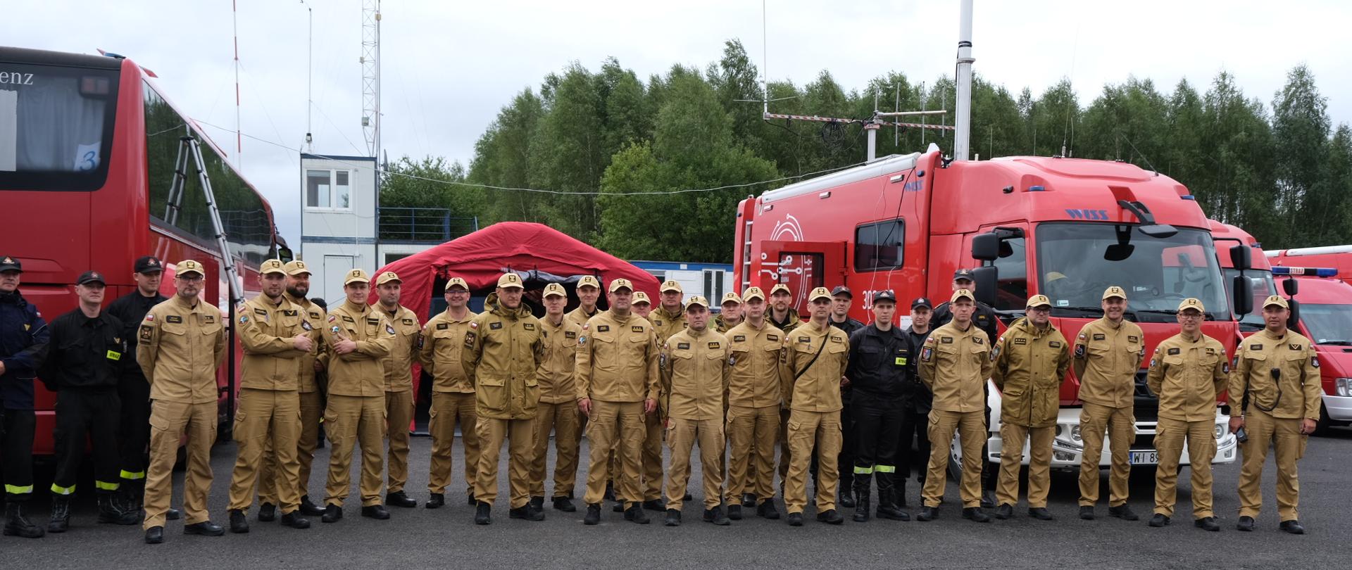 Grupa strażaków stoi na tle samochodów i namiotu PSP