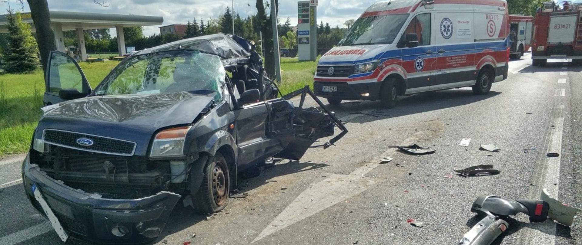 Widok na rozbity samochód osobowy marki Ford, na drugim planie ambulans. Na jezdni rozrzucone części rozbitego samochodu.