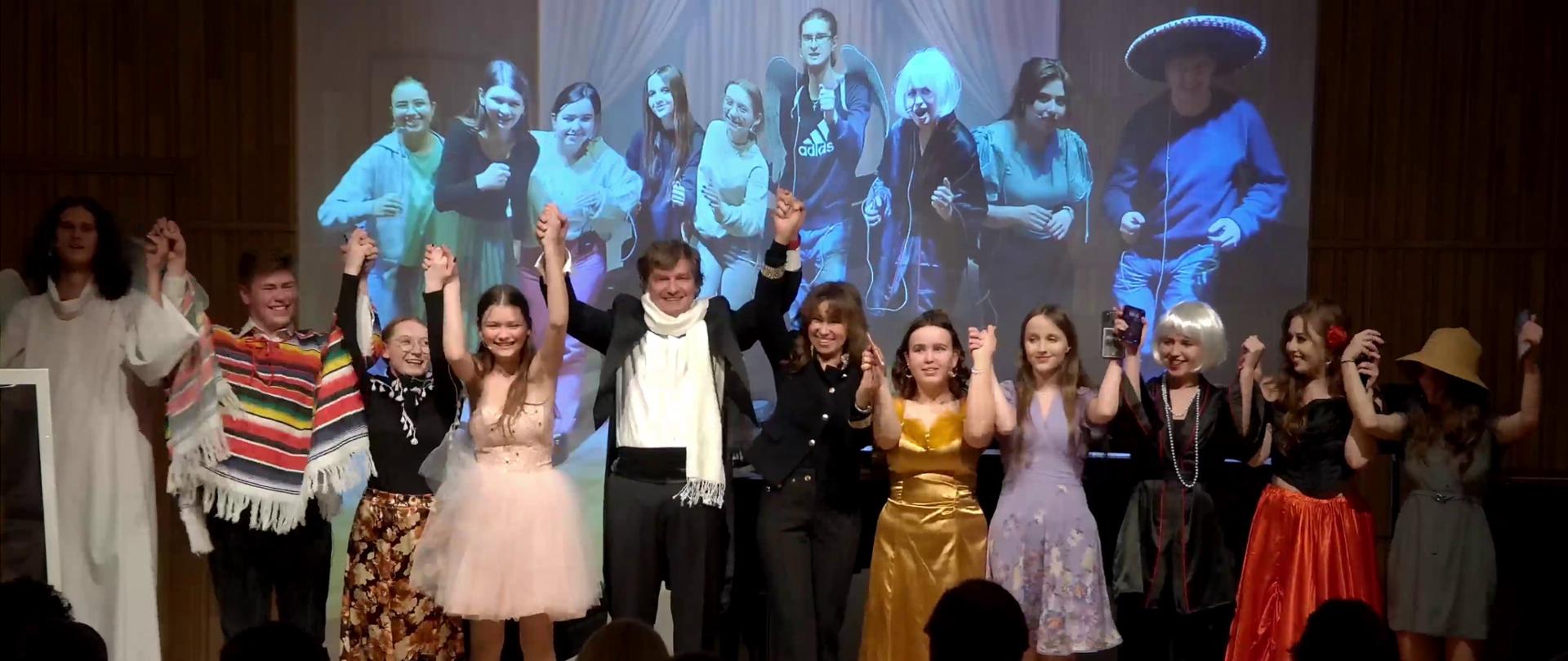 Zdjęcie. Na estradzie sali koncertowej stoją z uniesionymi rękoma wykonawcy musicalu "casting". Przed nimi stoją widzowie. W tle zdjęcie wykonawców z próby.