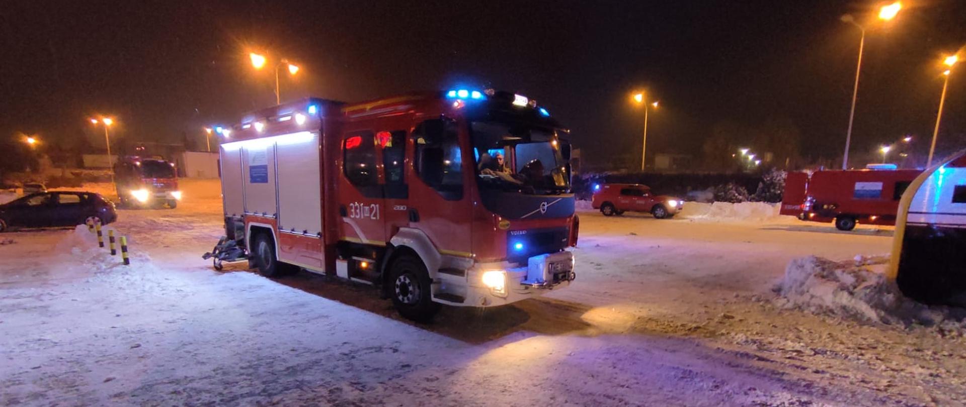 Na zdjęciu widać pojazdy ratowniczo – gaśnicze znajdujące się na parkingu przed obiektem, w którym doszło do pożaru. Pojazdy w kolorze czerwonym z włączonymi niebieskimi światłami sygnalizacji alarmowej. Parking pokrywa warstwa śniegu. Zdjęcie wykonane w porze nocnej. 