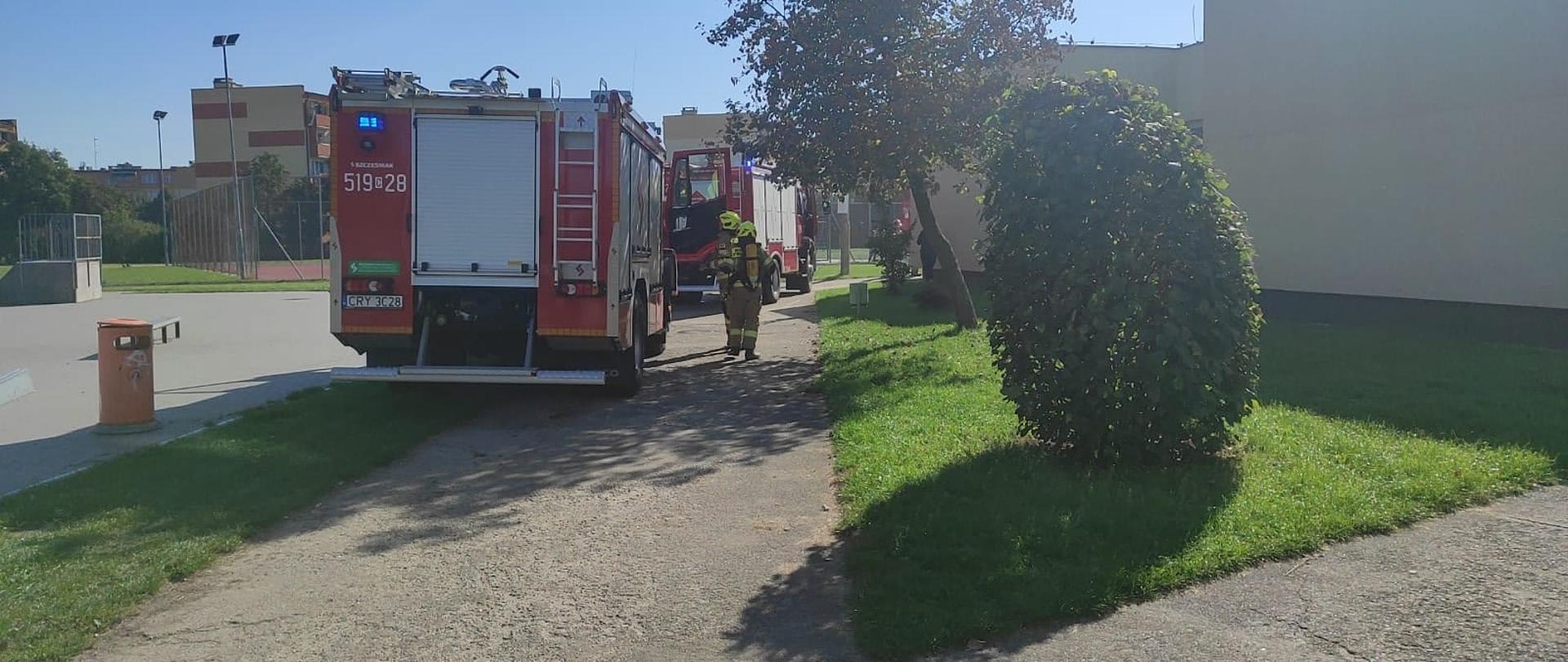 Dwa samochody pożarnicze stoją tyłem na boisku szkoły podstawowej numer 3. Obok samochodów po prawej stronie widać dwóch strażaków w pełnym uzbrojeniu.