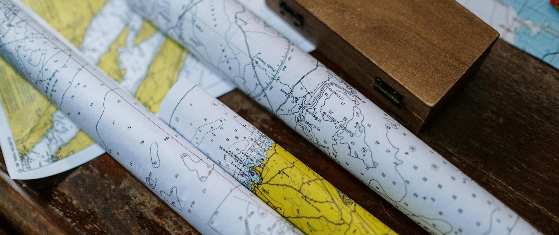 Zdjęcie prezentuje stół z kilkoma zwiniętymi mapa i pudełkiem najprawdopodobniej z kompasem - Zdjęcie dodane przez cottonbro studio: https://www.pexels.com/pl-pl/zdjecie/biale-i-brazowe-patyczki-drewniane-5302807/