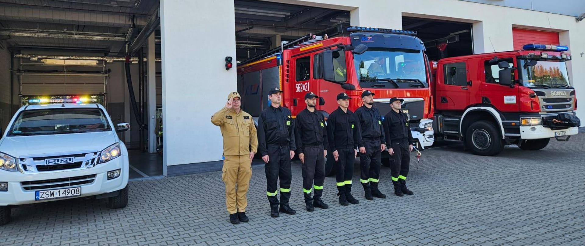 Strażacy salutują. Stoją w szeregu przed samochodami pożarniczymi. W tle budynek Jednostki Ratowniczo-Gaśniczej