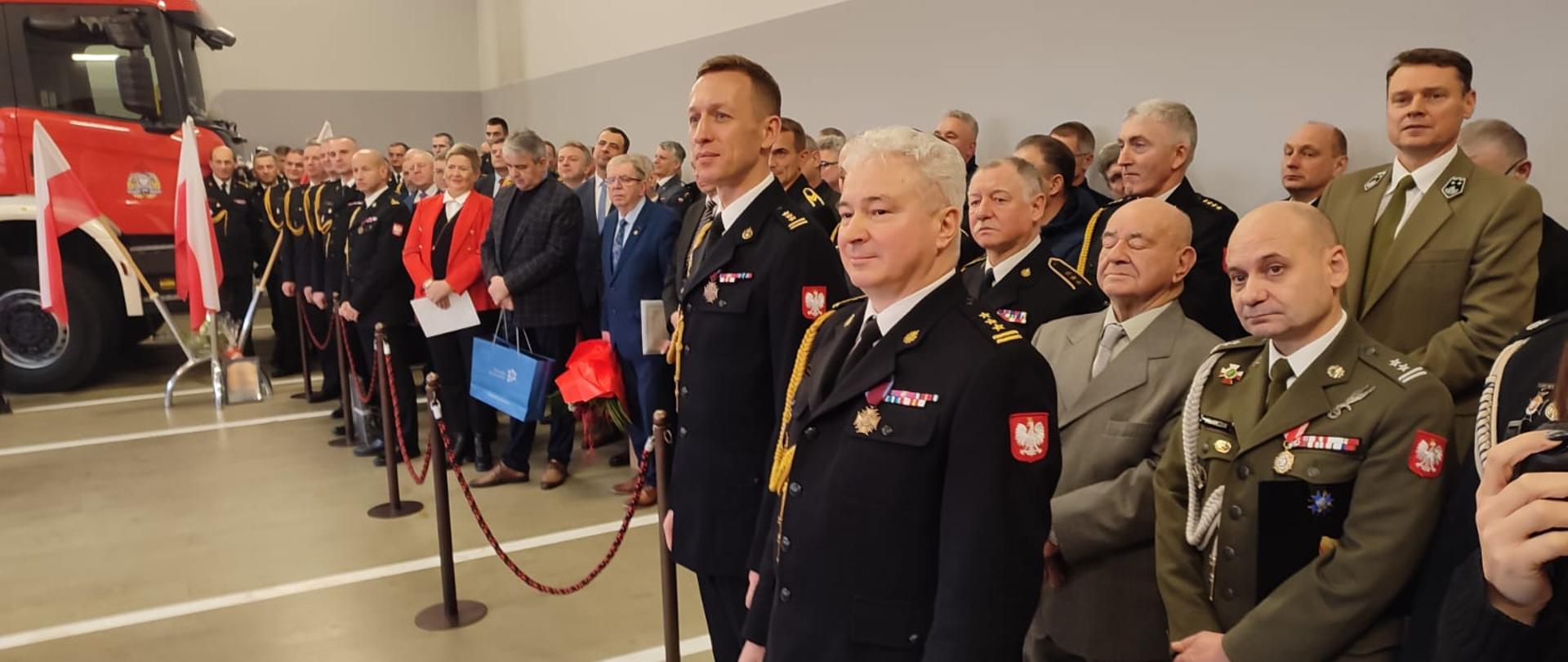 grupa ludzi - strażaków, zołnierzy, stoją w pomieszczeniu, na pierwszym planie dwóch strażaków w mundurach ze sznurem