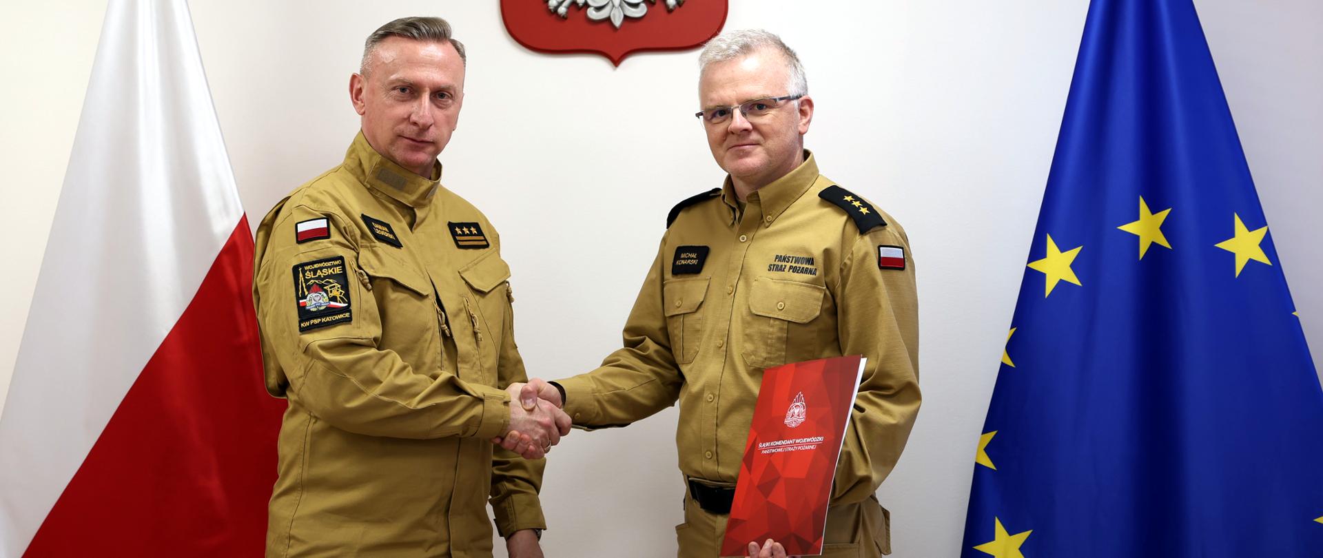 Na zdjęciu widać dwóch mężczyzn stojących obok siebie w pomieszczeniu. Obaj mają na sobie jednolite, piaskowo mundury służbowe państwowej straży pożarnej - z długimi rękawami, pagonami na ramionach oraz naszywkami na piersi i rękawach. Mężczyźni stoją przodem do siebie i podają sobie dłonie w geście uścisku. Po lewej stoi śląski komendant wojewódzki st. bryg. Damian Legierski trzyma ręce swobodnie, natomiast ten po prawej stoi mianowany naczelnik wydziału ochrony ludności kpt. Michał Konarski, który trzyma w lewej ręce czerwoną teczkę z aktem mianowania. Za nimi, na ścianie, centralnie umieszczony jest herb Polski – biały orzeł na czerwonym tle. Po lewej stronie kadru stoi pionowo flaga Polski, biała u góry i czerwona u dołu. Po prawej stronie znajduje się flaga Unii Europejskiej, niebieska z okręgiem żółtych gwiazd.