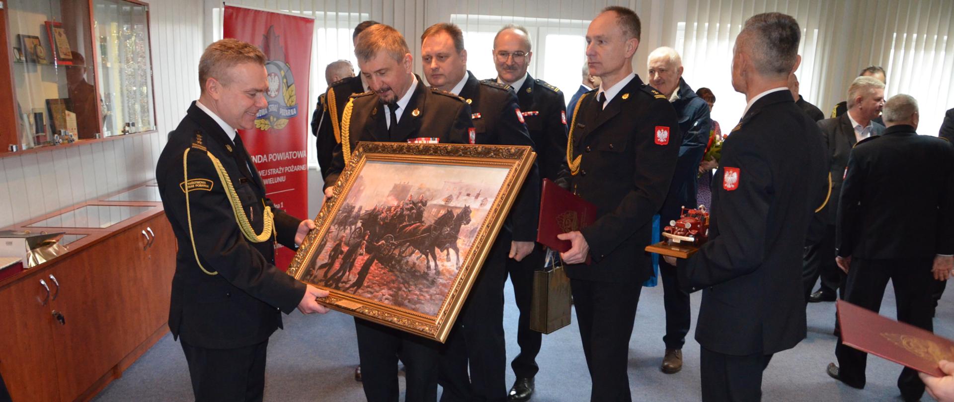 st. bryg. Piotr Cały otrzymuje od st. bryg. Pawła Malinowskiego obraz przedstawiający wyjeżdzających do akcji strażaków w wozie konnym