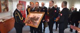 st. bryg. Piotr Cały otrzymuje od st. bryg. Pawła Malinowskiego obraz przedstawiający wyjeżdzających do akcji strażaków w wozie konnym