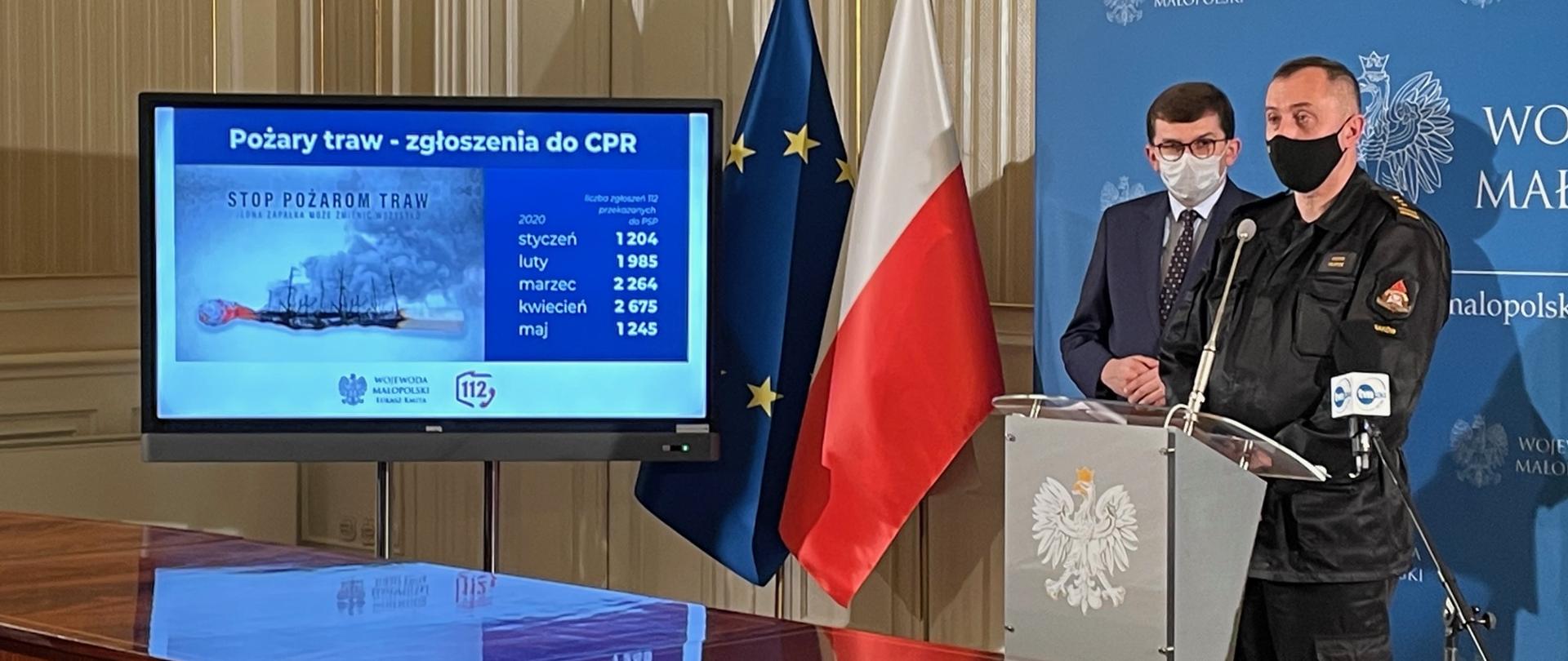 Podczas briefingu prasowego dotyczącego przełączania numeru alarmowego 998 do Centrum Powiadamiania Ratunkowego przemawia małopolski komendant wojewódzki Państwowej Straży Pożarnej st. bryg. Piotr Filipek po jego prawej stronie stoi wojewoda małopolski Łukasz Kmita. Na telewizorze wyświetla się infografika mówiąca wzroście liczbie połączeń alarmowych w związku z pożarami traw.