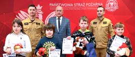 Laureaci turnieju na tle ścianki z koloru czerwonego w obecności dwóch strażaków oraz pana w garniturze