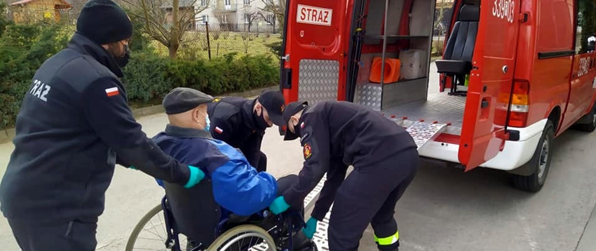 Na zdjęciu widoczny mężczyzna w podeszłym wieku na wózku, którego strażacy przemieszczają do strażackiego czerownego busa. Strażaków jest trzech, ubranych w umundurowaniu koszarowe, mają na sobie maseczki i rękawiczki. Bus stoi zaparkowany tyłem, tyłem i z profilu zwróceni są również strażacy i mężczyzna.