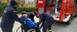 Na zdjęciu widoczny mężczyzna w podeszłym wieku na wózku, którego strażacy przemieszczają do strażackiego czerownego busa. Strażaków jest trzech, ubranych w umundurowaniu koszarowe, mają na sobie maseczki i rękawiczki. Bus stoi zaparkowany tyłem, tyłem i z profilu zwróceni są również strażacy i mężczyzna.