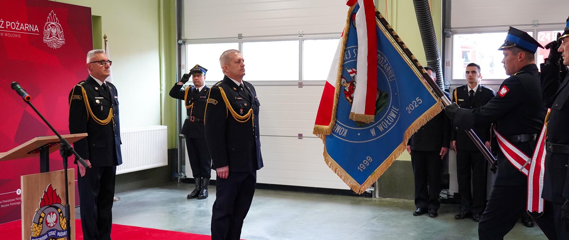 Zdjęcie przedstawia uroczysty moment oficjalnej ceremonii Państwowej Straży Pożarnej wewnątrz garażu jednostki. Nowo powołany komendant powiatowy PSP w Wołowie stoi na baczność na jaskrawoczerwonym dywanie reprezentacyjnym, naprzeciwko pocztu sztandarowego wprowadzającego sztandar jednostki. W uroczystości biorą udział inni wyżsi oficerowie oraz zaproszeni goście. Nowo powołany komendant w centrum kadru stoi na baczność na środku czerwonego dywanu, zwrócony profilem w prawą stronę. Ma podniesioną głowę i patrzy z powagą na prezentowany przed nim sztandar. Nosi ciemnogranatowy mundur wyjściowy ze złotym sznurem galowym na prawym ramieniu, dystynkcjami na pagonach oraz czarne, eleganckie półbuty. W tyle stoi na baczność na skraju dywanu Zastępca Dolnośląskiego Komendanta Wojewódzkiego PSP, tuż obok drewnianej mównicy z dużym, kolorowym logo Państwowej Straży Pożarnej. Nosi ciemnogranatowy mundur ze sznurem galowym i okularami w ciemnych oprawkach. Obserwuje ceremonię. Strażak niosący sztandar widoczny w dynamicznej postawie od pasa w dół oraz z profilu, trzyma oburącz pochylone pod kątem drzewce sztandaru. Przez ramię ma przewieszoną biało-czerwoną szarfę pocztową. Nosi wysokie, czarne buty taktyczne ze sznurowaniem. Asysta sztandaru: Obok niego, tyłem do obiektywu, stoi kolejny mundurowy w czapce rogatywce, salutujący prawą dłonią.