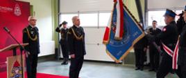Zdjęcie przedstawia uroczysty moment oficjalnej ceremonii Państwowej Straży Pożarnej wewnątrz garażu jednostki. Nowo powołany komendant powiatowy PSP w Wołowie stoi na baczność na jaskrawoczerwonym dywanie reprezentacyjnym, naprzeciwko pocztu sztandarowego wprowadzającego sztandar jednostki. W uroczystości biorą udział inni wyżsi oficerowie oraz zaproszeni goście. Nowo powołany komendant w centrum kadru stoi na baczność na środku czerwonego dywanu, zwrócony profilem w prawą stronę. Ma podniesioną głowę i patrzy z powagą na prezentowany przed nim sztandar. Nosi ciemnogranatowy mundur wyjściowy ze złotym sznurem galowym na prawym ramieniu, dystynkcjami na pagonach oraz czarne, eleganckie półbuty. W tyle stoi na baczność na skraju dywanu Zastępca Dolnośląskiego Komendanta Wojewódzkiego PSP, tuż obok drewnianej mównicy z dużym, kolorowym logo Państwowej Straży Pożarnej. Nosi ciemnogranatowy mundur ze sznurem galowym i okularami w ciemnych oprawkach. Obserwuje ceremonię. Strażak niosący sztandar widoczny w dynamicznej postawie od pasa w dół oraz z profilu, trzyma oburącz pochylone pod kątem drzewce sztandaru. Przez ramię ma przewieszoną biało-czerwoną szarfę pocztową. Nosi wysokie, czarne buty taktyczne ze sznurowaniem. Asysta sztandaru: Obok niego, tyłem do obiektywu, stoi kolejny mundurowy w czapce rogatywce, salutujący prawą dłonią.