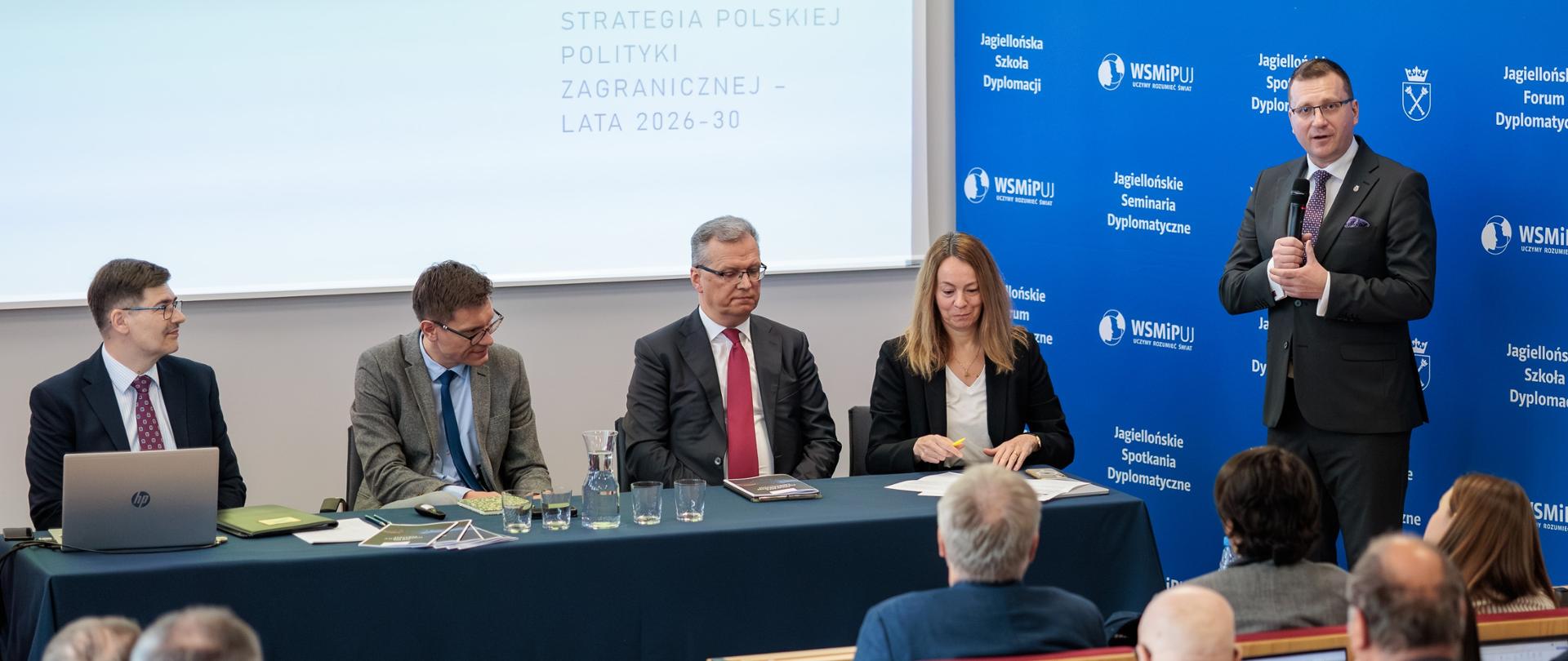 The photo shows five people: three men and a woman sitting at a long table. One man is standing. They are addressing the audience. Behind them is a sign that reads “Polish Foreign Policy Strategy 2026–2023.” Next to them is a blue backdrop with white lettering that reads “Jagiellonian Diplomatic Meetings.”