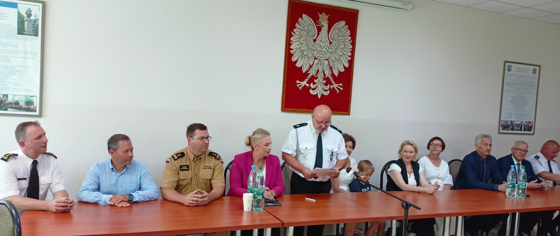grupa osób , w tym strażacy siedzą za stołem, strażak w białej koszuli i krawacie, strażak OSP stoi i przemawia do mikrofonu, za nimi biała ściana na której wisi godło polski - biały orzeł na czerwonym tle