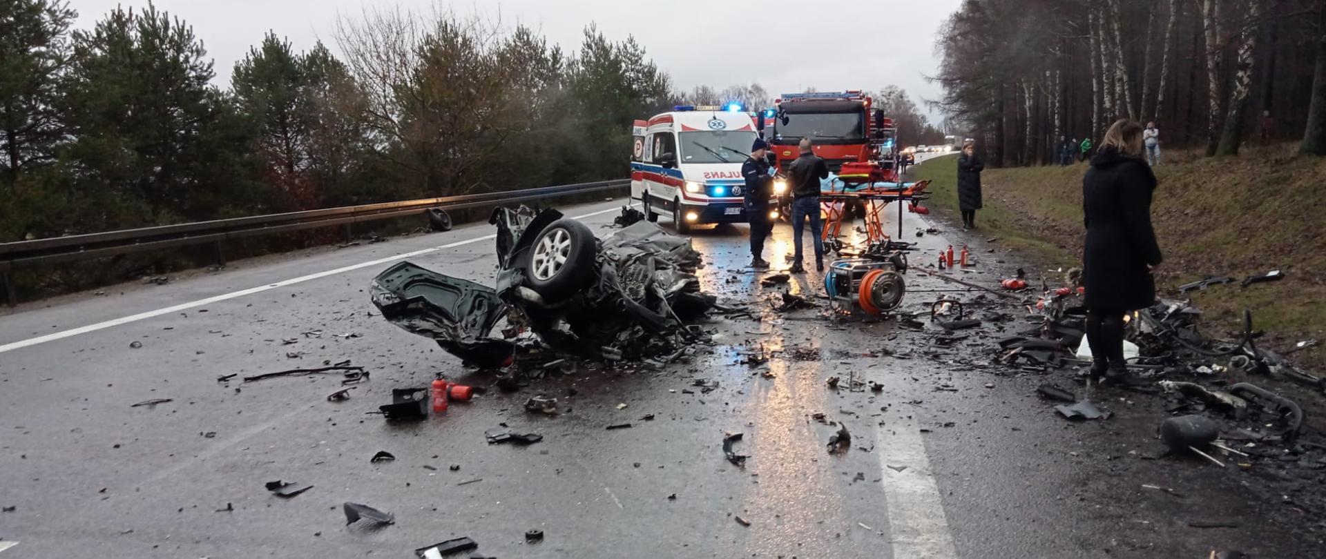 Zdjęcie przedstawia porozrzucane elementy karoserii pojazdów oraz część rozerwanego pojazdu. W tle widoczna karetka pogotowia, samochód pożarniczy oraz zestaw hydrauliczny.