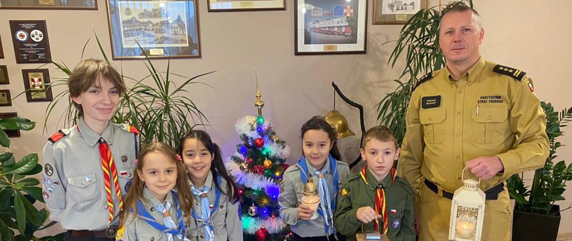 Cztery zuszki w mundurach wraz z drużynową przekazują komendantowi powiatowemu światełko. Za nimi widać choinkę i obrazy na ścianach.