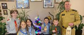 Cztery zuszki w mundurach wraz z drużynową przekazują komendantowi powiatowemu światełko. Za nimi widać choinkę i obrazy na ścianach.