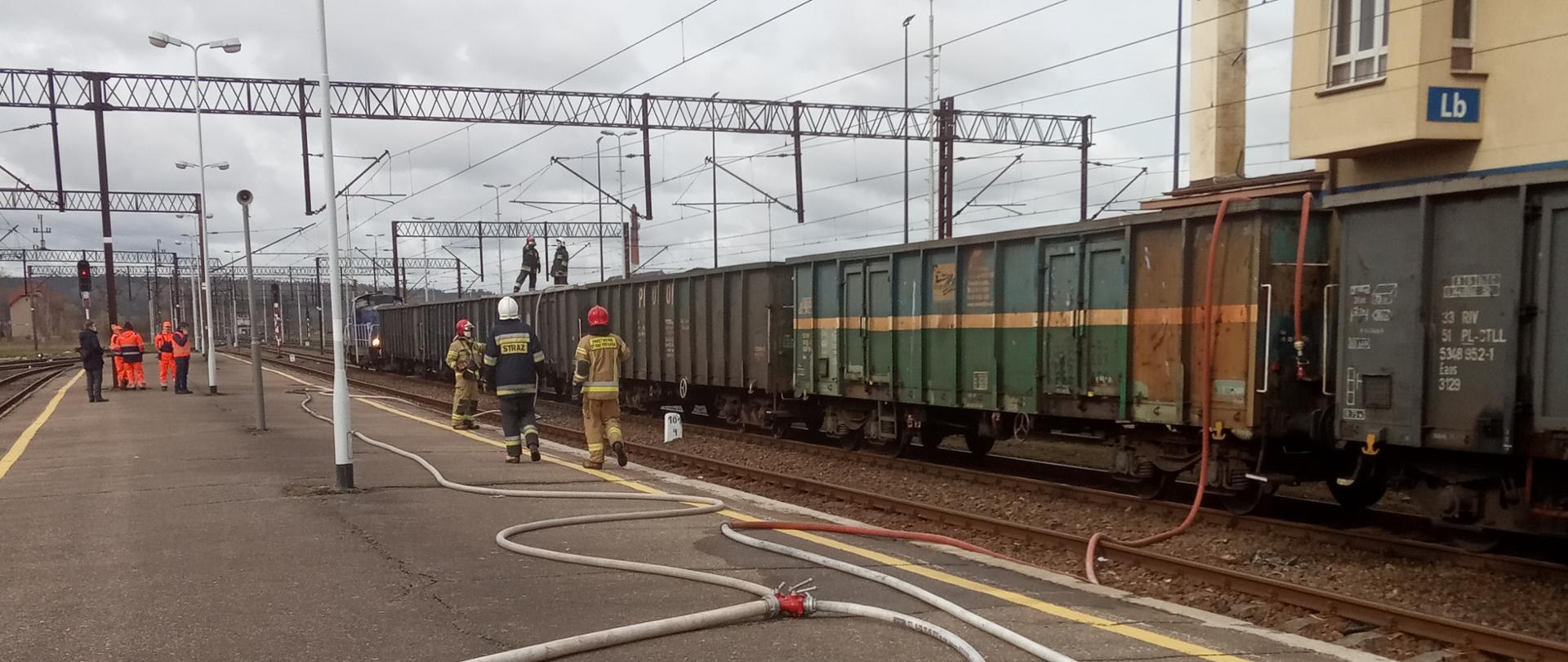 Na zdjęciu widoczny jest peron na którym stacjonuje skład pociągu towarowego. Przez peron przebiega linia główna zakończona rozdzielaczem oraz dwie linie gaśnicze prowadzące do wagonów. W wagonach znajdują się strażacy operujący prądami gaśniczymi. W dalszej części peronu widoczni są przedstawiciele polskich lini kolejowych.