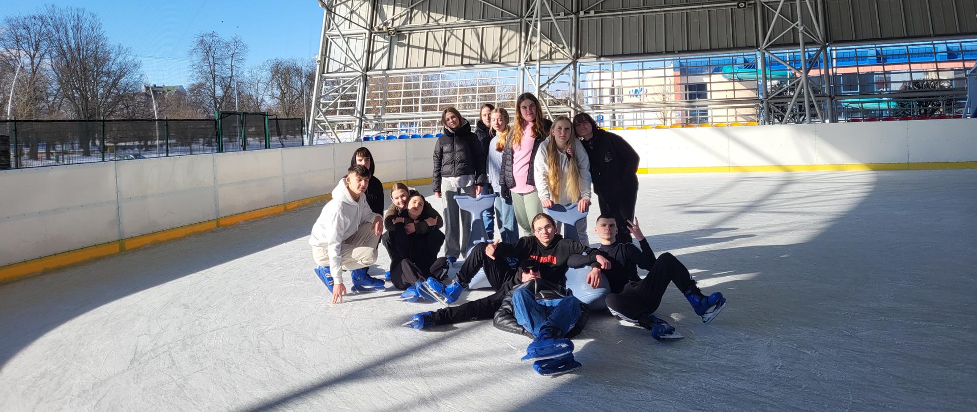 Grupa młodzieży na lodowisku, kilka osób siedzi na lodzie, kiloka stoi
