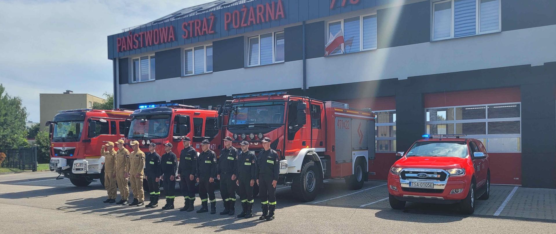 Na zbiórce przed remizą znajdują się strażacy w mundurach. W tle samochody bojowe.