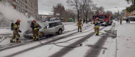 Zdjęcie przedstawia strażaków w trakcie działań przy samochodzie osobowym koloru srebrnego. Samochód ma otwartą maskę, z komory silnika wydobywa się dym. Na zdjęciu widocznych jest 4 strażaków w jasnych piaskowych mundurach w czerwonych hełmach. W tle widoczne samochody pożarnicze koloru czerwonego. Zdarzenie ma miejsce na zaśnieżonej jezdni.