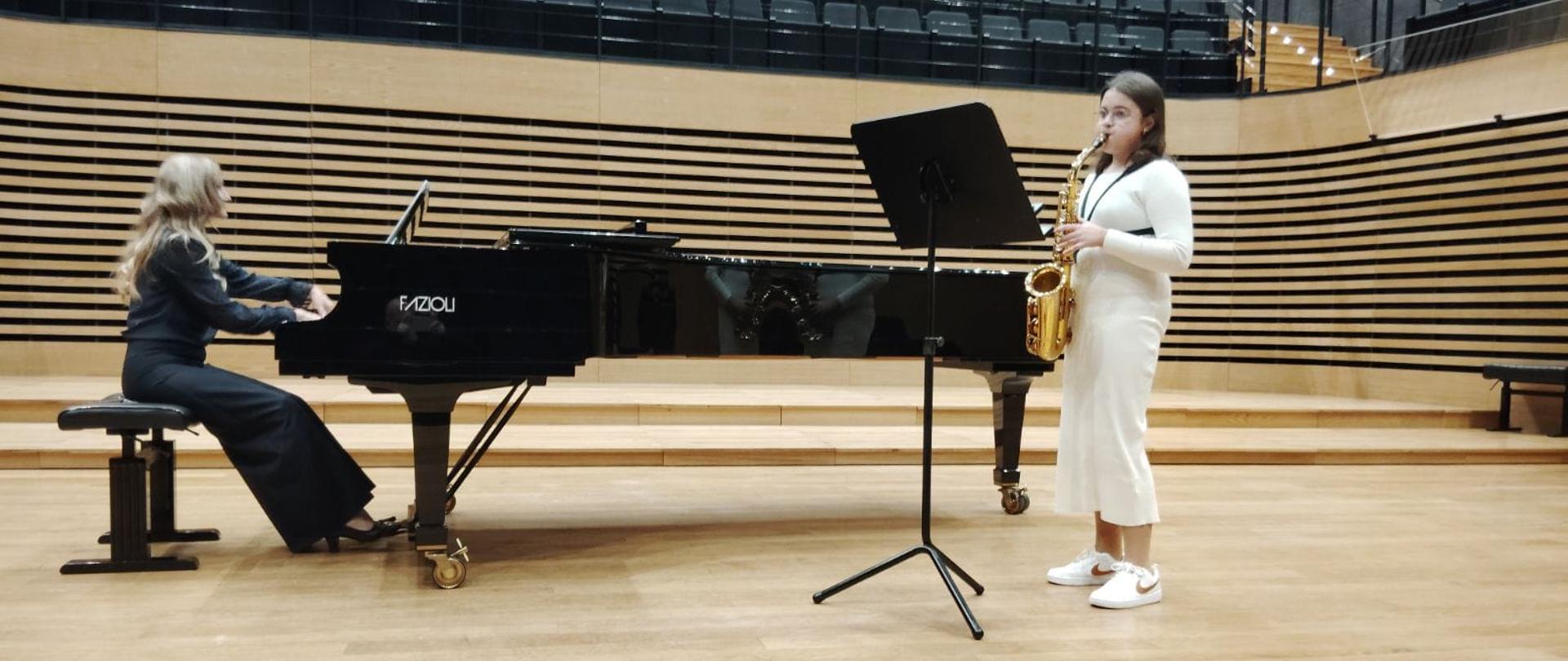 Dziewczynka gra na saksofonie, kobieta akompaniuje na fortepianie.