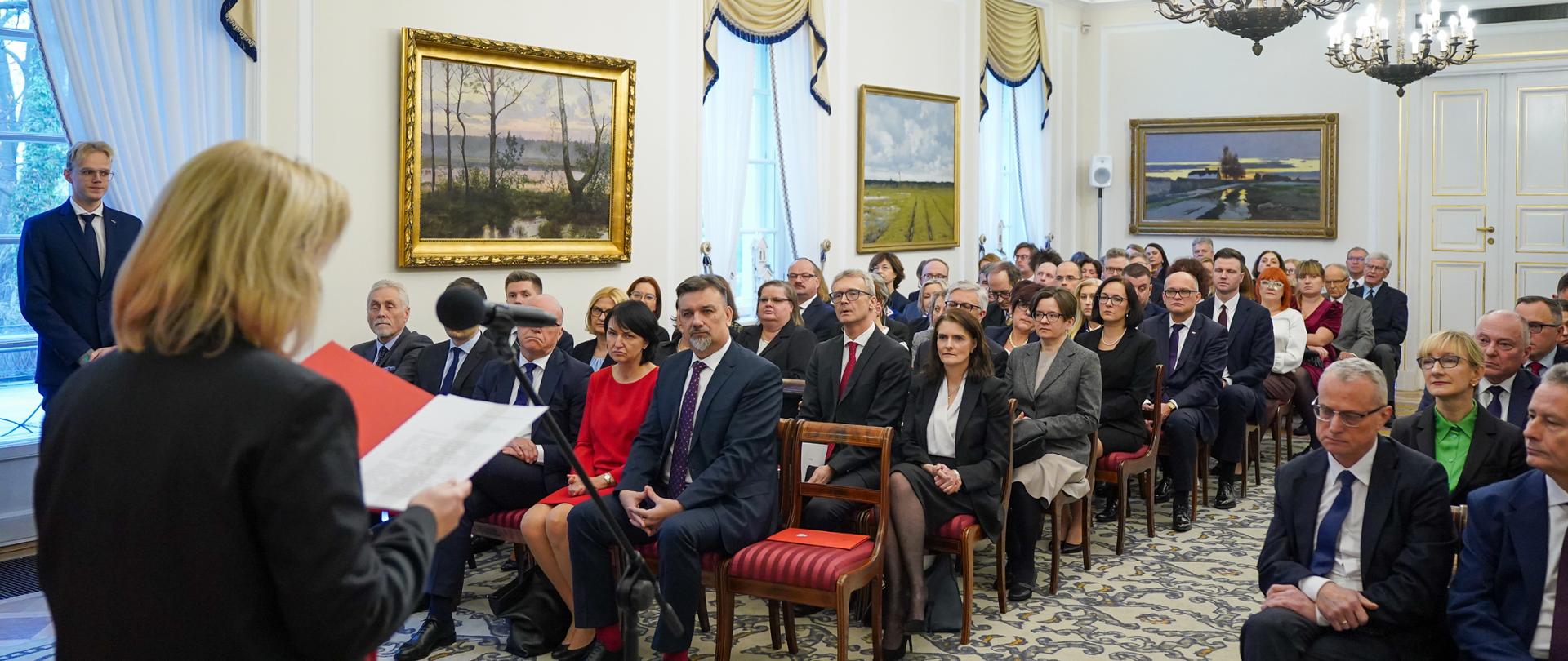 Na zdjęciu jest kilkadziesiąt osób. Siedzą na krzesłach w dużej sali. Przed nimi stoi kobieta w ciemnym ubraniu. Mówi do mikrofonu.

