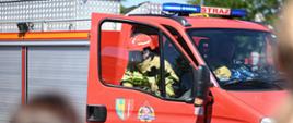Strażak w kabinie czerwonego wozu strażackiego z włączonym sygnałem świetlnym podczas pokazu na pikniku bezpieczeństwa.
