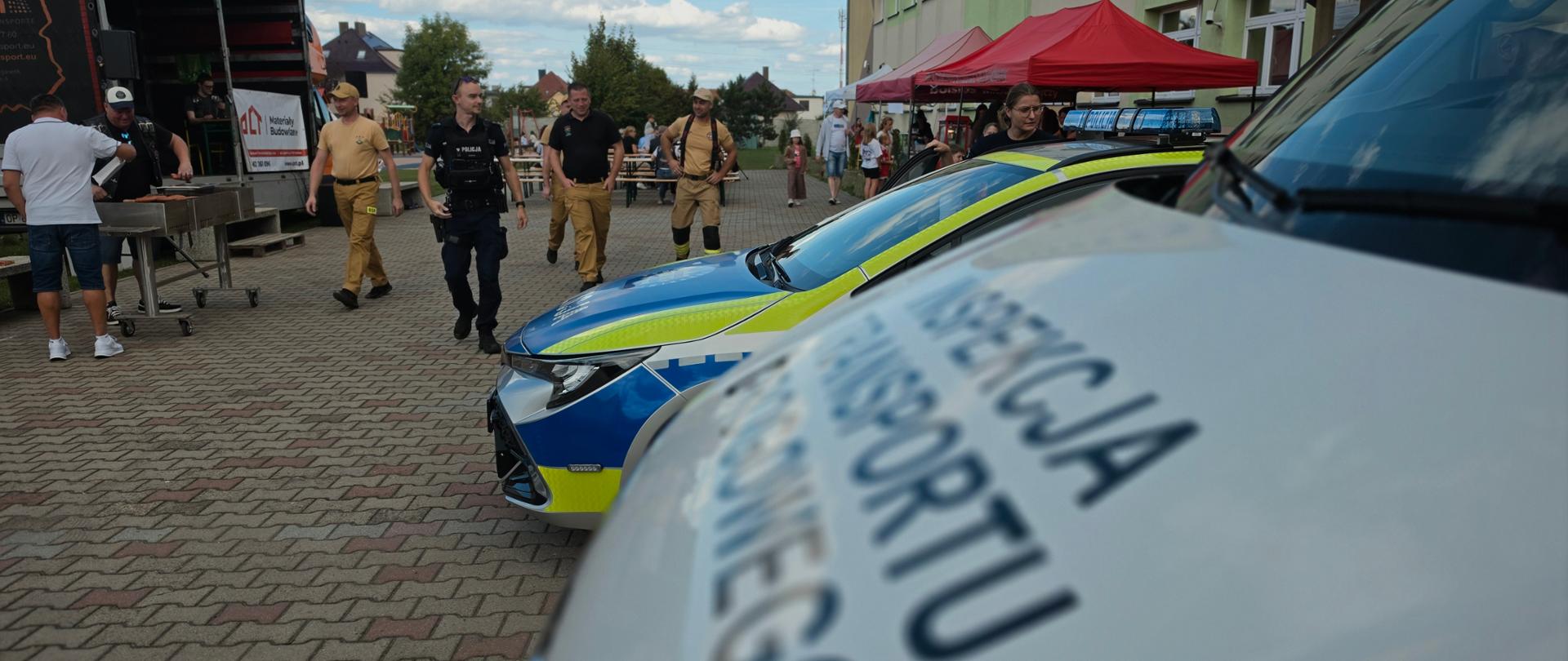 Na pierwszym planie widoczny jest fragment oznakowanego pojazdu Inspekcji Transportu Drogowego z dużym napisem „Inspekcja Transportu Drogowego”. Tuż za nim stoi oznakowany radiowóz Policji z niebieską belką świetlną na dachu. W głębi, na placu przed szkołą, poruszają się funkcjonariusze oraz inni uczestnicy wydarzenia. W tle widoczne są namioty, stoły oraz budynek szkoły.