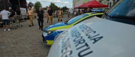 Na pierwszym planie widoczny jest fragment oznakowanego pojazdu Inspekcji Transportu Drogowego z dużym napisem „Inspekcja Transportu Drogowego”. Tuż za nim stoi oznakowany radiowóz Policji z niebieską belką świetlną na dachu. W głębi, na placu przed szkołą, poruszają się funkcjonariusze oraz inni uczestnicy wydarzenia. W tle widoczne są namioty, stoły oraz budynek szkoły.