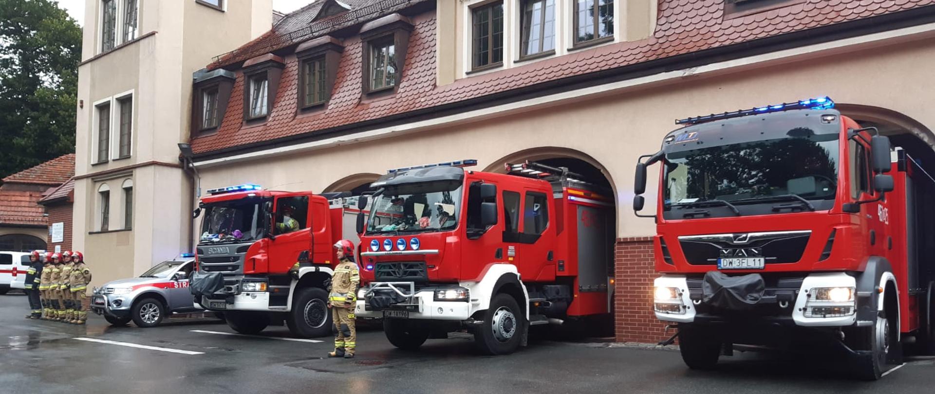 Cztery pojazdy ratowniczo-gaśnicze przed bramami wyjazdowymi z włączonymi sygnałami świetlnymi. Przed samochodami w szeregu stoją strażacy ubrani w umundurowanie specjalne.